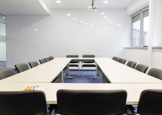 Ustawienie sali konferencyjnej z długim stołem i rzędami krzeseł po obu stronach, z projektorem na suficie i materiałami piśmiennymi na stole.